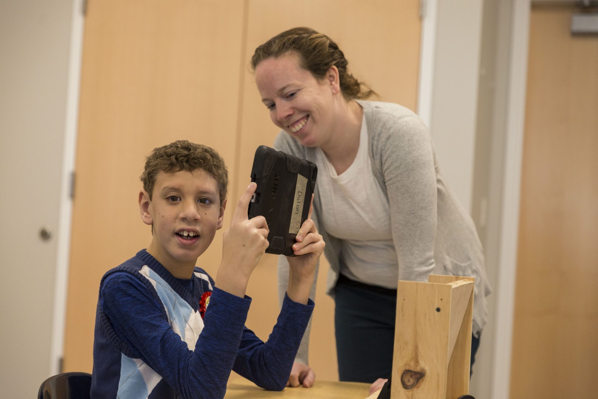 A young boy sits at his desk with a tablet and it smiling. An ABA Specialist is standing next to him also smiling.